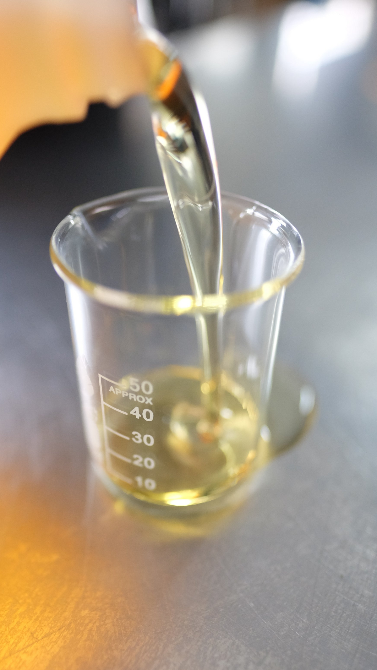 A clear glass beaker with measurement markings on a metallic gray surface, with a golden-yellow liquid being poured into it from an out-of-focus hand or container.